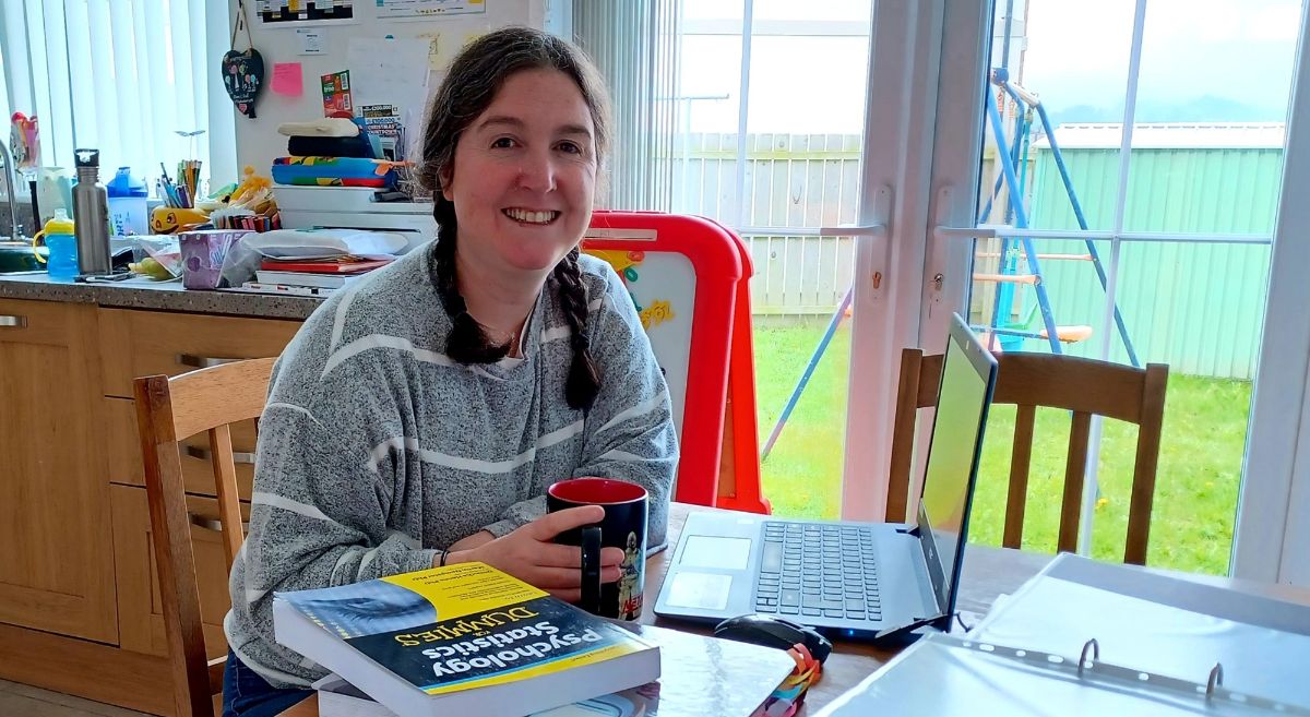 Jenny Greer, female, smiling to camera with brown hair in plaits, wearing grey and white sweater, sitting at kitchen table with coffee cup and text books and files laid out before her.